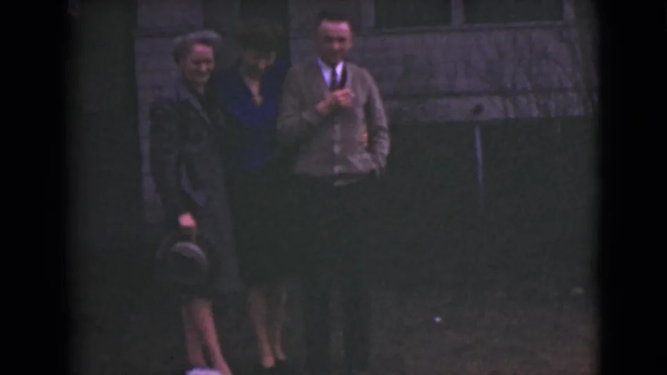 A woman in a dark coat holds a small hat while standing with another woman and a man in 1940s clothing outside in Alaska.