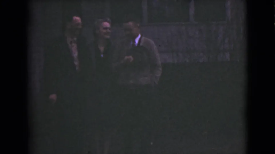 In metadata-supported 1946 Alaska footage, two men and a woman stand together in low light near a dark building facade.