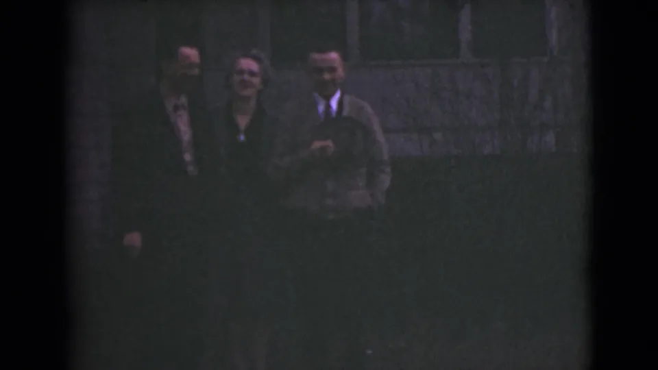 The dim 8mm frame shows the same three adults in formal clothing, standing on dark ground with a building behind them.