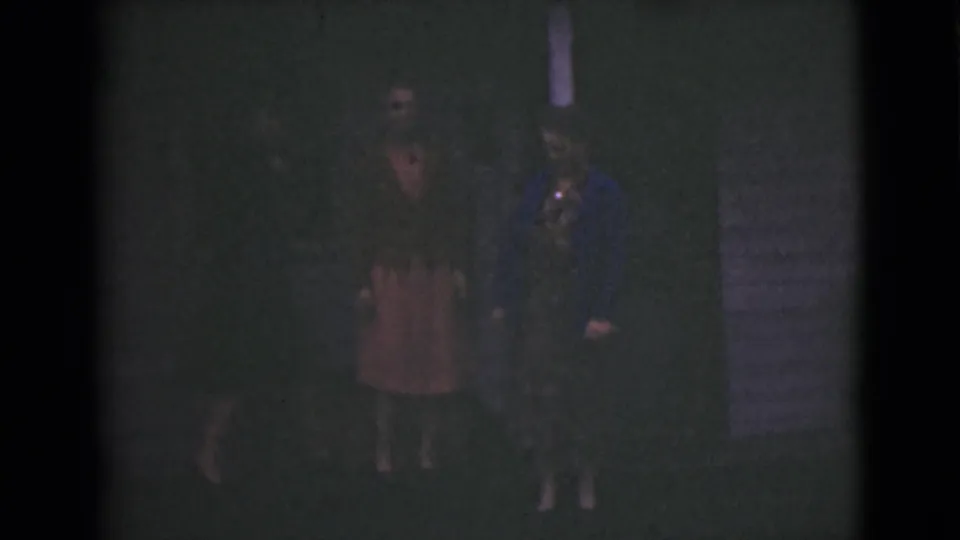 A grainy 8mm frame shows two women by an exterior wall at night, with clothing and faces faint in the dark exposure.