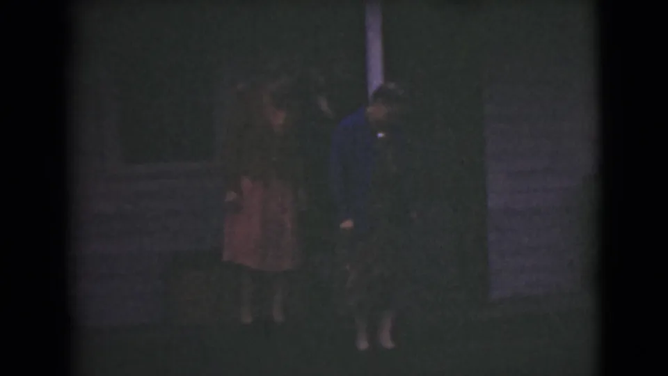 The final frame shows two adult women in dresses beside a building exterior, filmed at night on dark 8mm footage.
