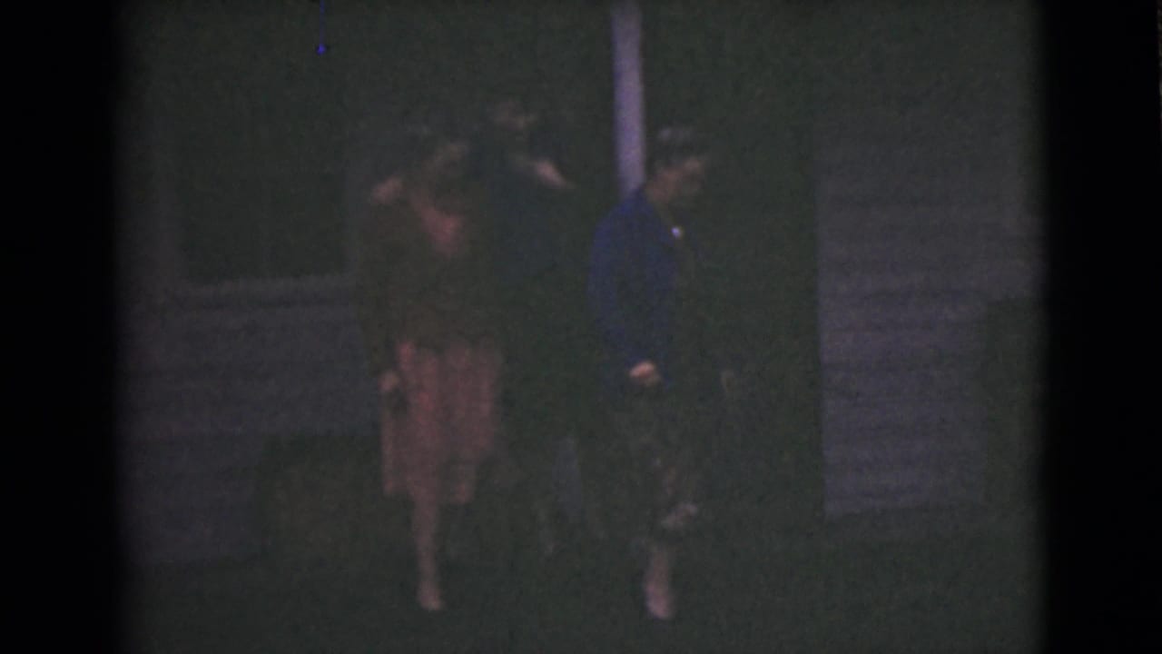 Three women walk on grass outside a dark house at night.