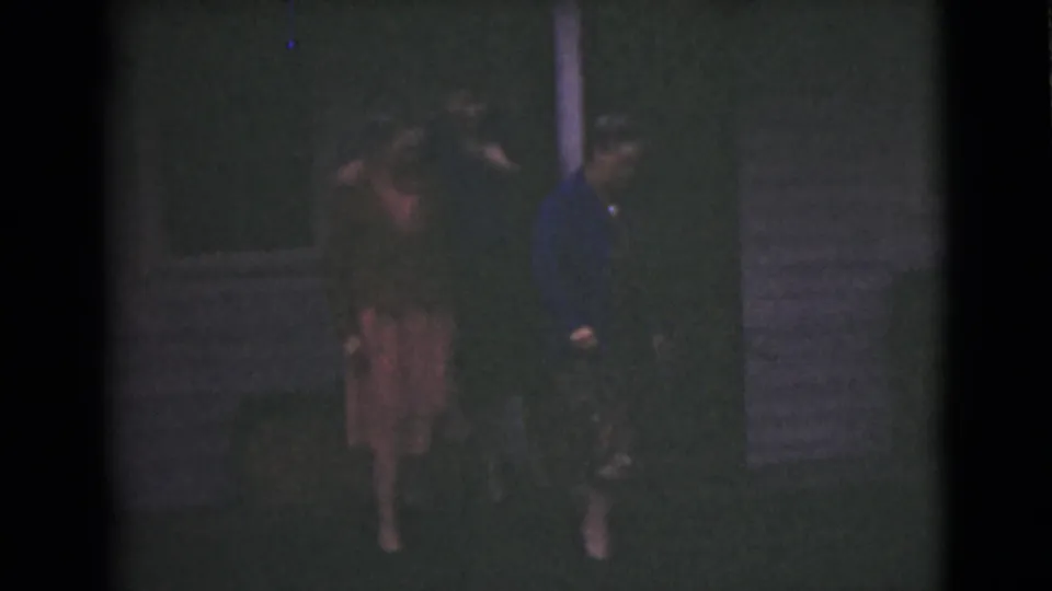 In 1946 Alaska home-movie footage, three women walk together outside a dim house, with dresses and jackets partly visible.