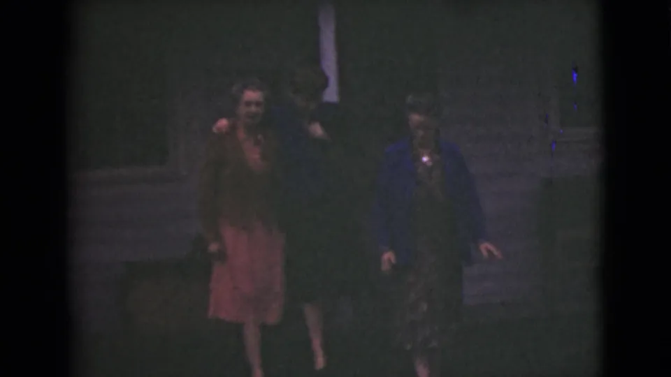 Three women in mid-century clothing walk close together outside a house in dim 1946 Alaska home-movie footage.