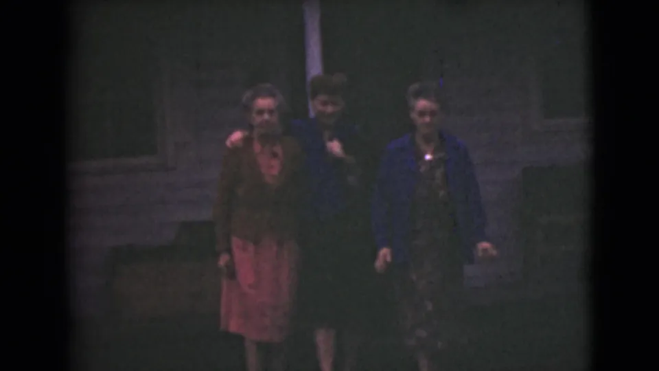 A woman in a red dress, a woman in a dark dress, and an older woman in a blue jacket appear outside a house at night.