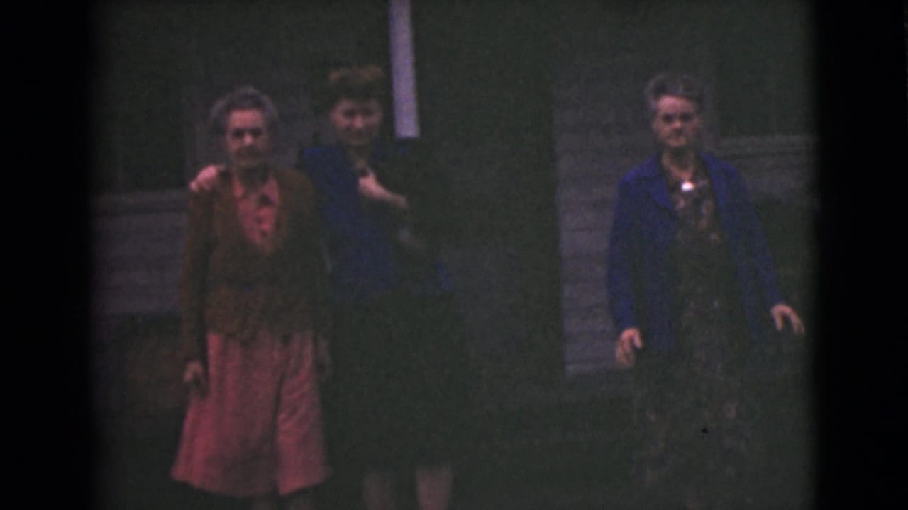 Three older women walk on grass outside a wooden building.