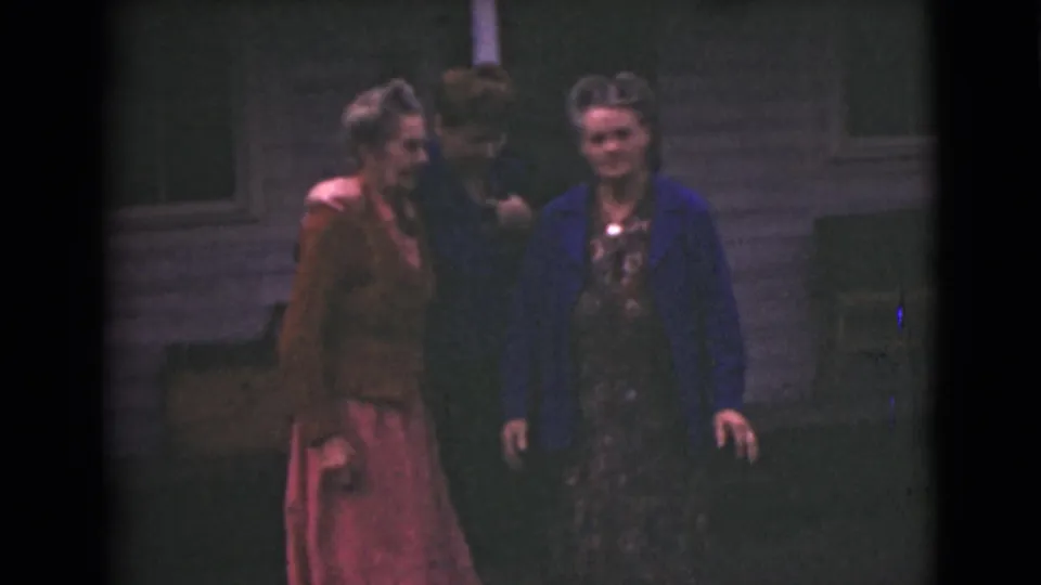 Two older women walk in front while a third follows closely, seen in soft 8mm color outside a wooden building in Alaska.