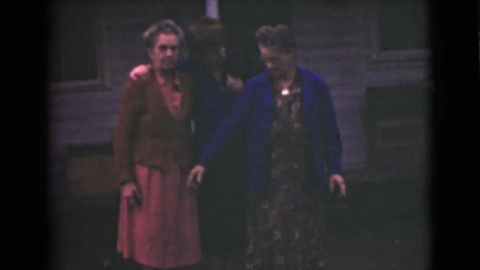 Three older women continue walking together outdoors in Alaska, framed by grass and a wooden building wall.