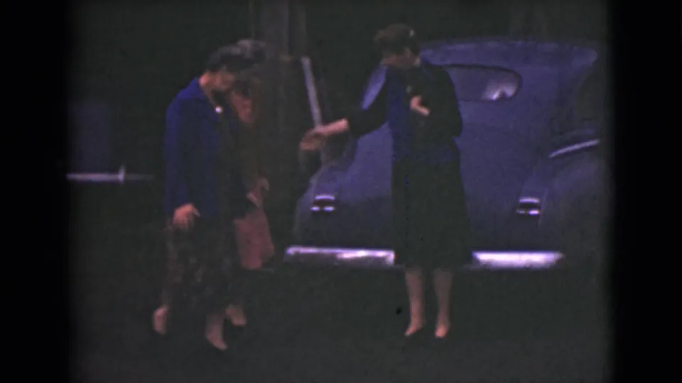 Three adult women in 1940s attire stand near the rear of a dark classic car, with one woman reaching toward another.