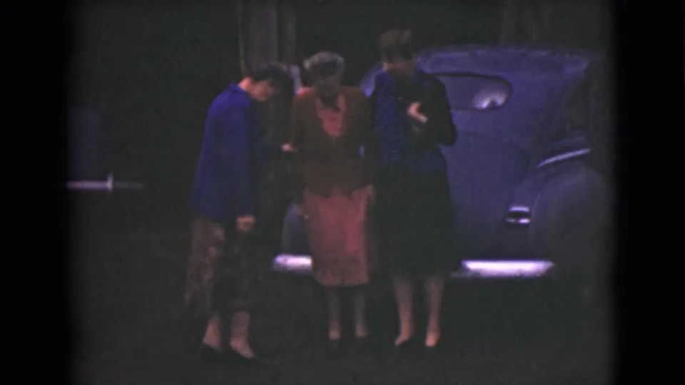 In the dim 1946 Alaska scene, three women stand close near a classic car as they adjust their stance before the photograph.
