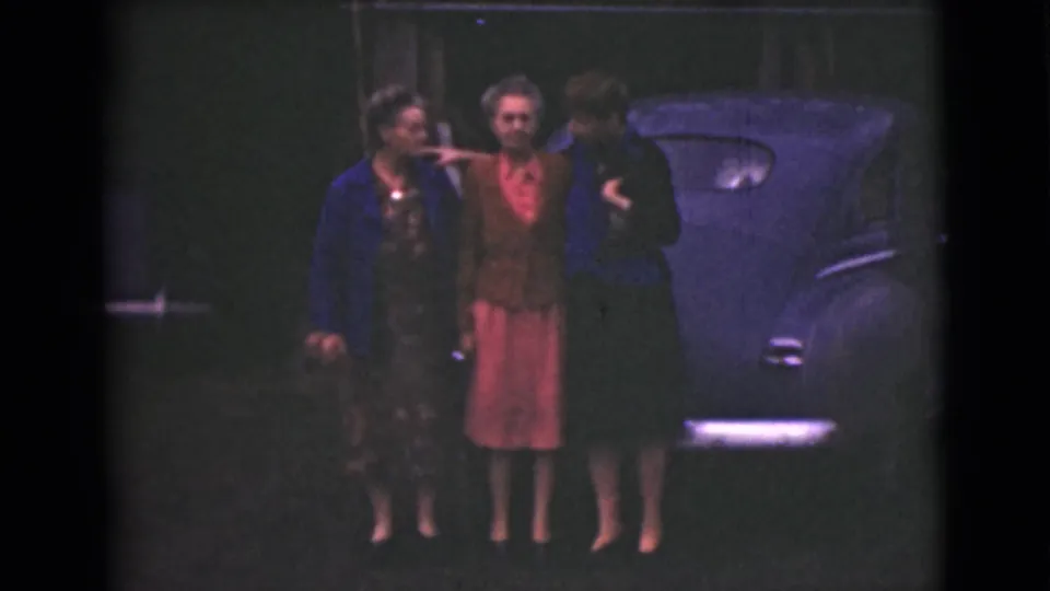 Three adult women pose arm in arm outdoors in Alaska in 1946, standing in front of a dark rounded car on 8mm film.