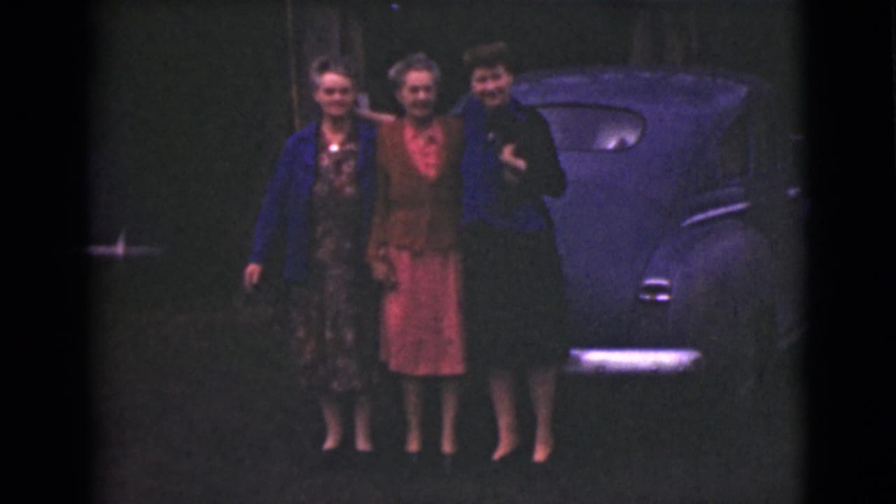 Three women pose beside a rounded blue car in a dim outdoor setting.