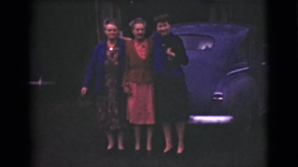 Three older women in dresses and jackets pose together beside a rounded blue car in 1946 Alaska home-movie footage.