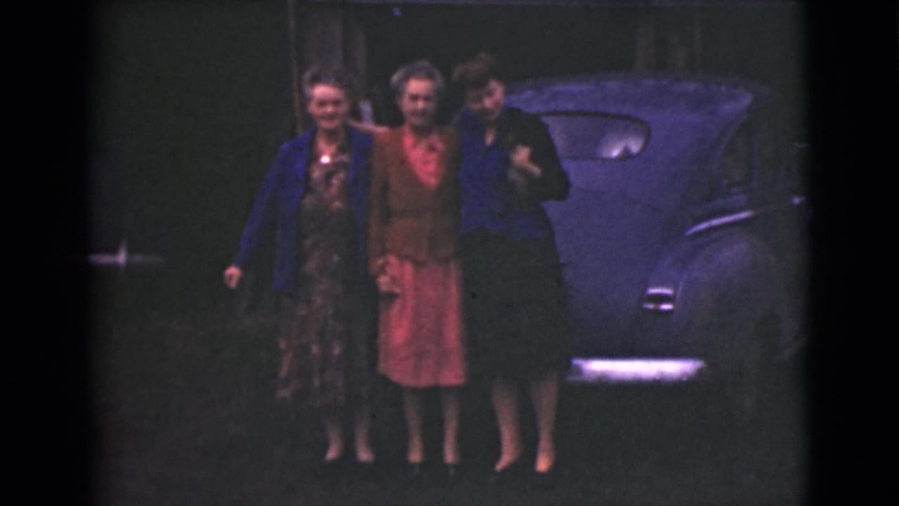 Three women continue posing near the rear side of a blue car.