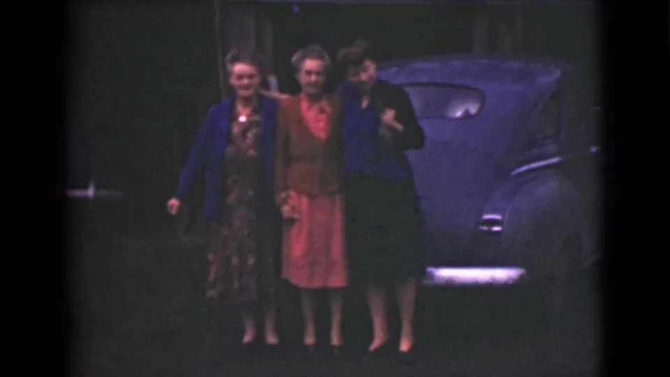 The women remain grouped beside the vintage blue car, smiling toward the camera in the Alaska home-movie shot.