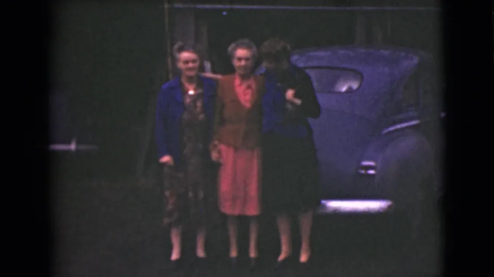 Three older women stand shoulder to shoulder in front of the rounded car, with muted color and soft 8mm texture.