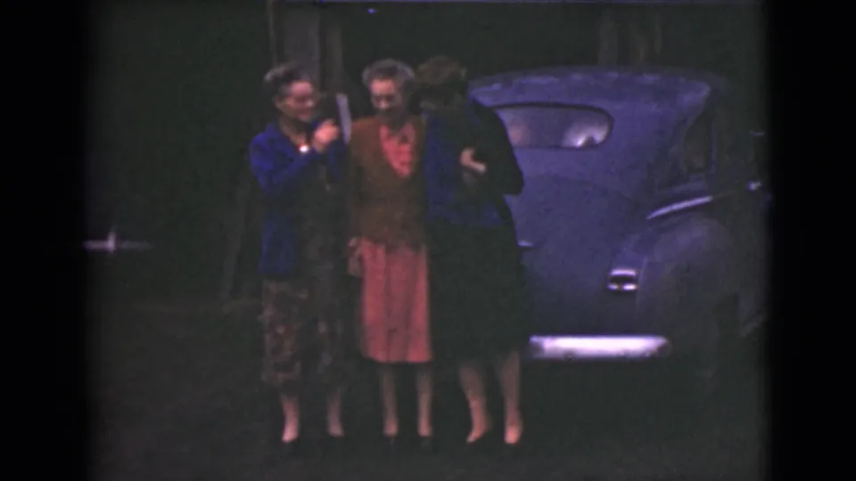 One woman appears to lift a small object as the three women laugh or smile beside the blue car in 1946 Alaska footage.