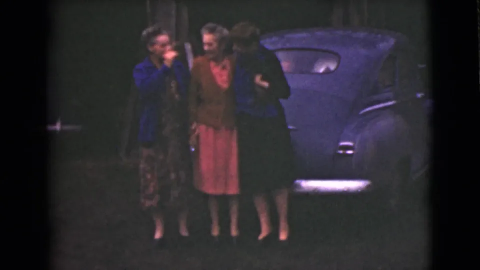 Three adult women in 1940s clothing stand beside a dark car at night or dusk in Alaska, according to clip metadata.