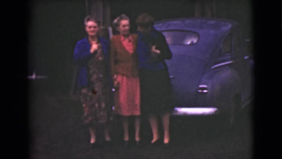 Three adult women appear to converse beside a parked dark car in a low-light 1946 Alaska home-movie frame.