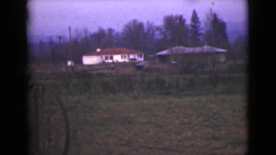 1946 Alaska home-movie frame with a white red-roofed house and nearby structure across open ground, framed by trees.