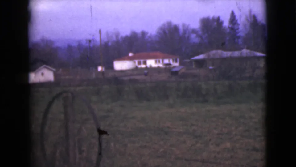 1946 Alaska 8mm frame showing a small pale shed, distant red-roofed house, fence post, and wooded background.
