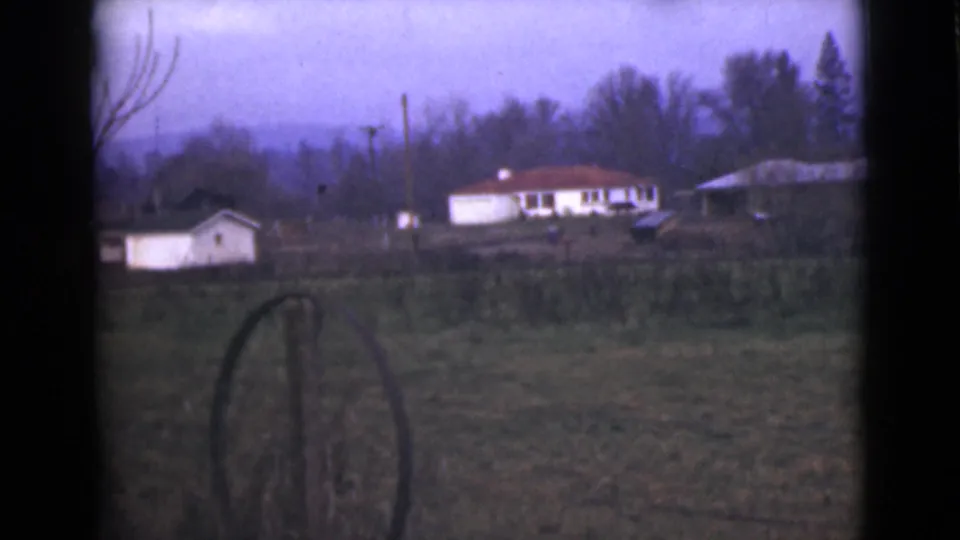 1946 Alaska home-movie frame with distant houses, a foreground fence post, open field, trees, and soft purple sky.