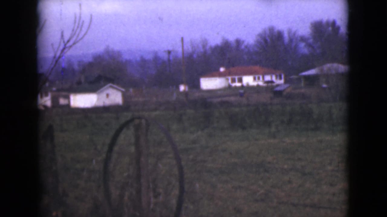 Rural field with fence posts, small white buildings, bare branches, and distant hills.