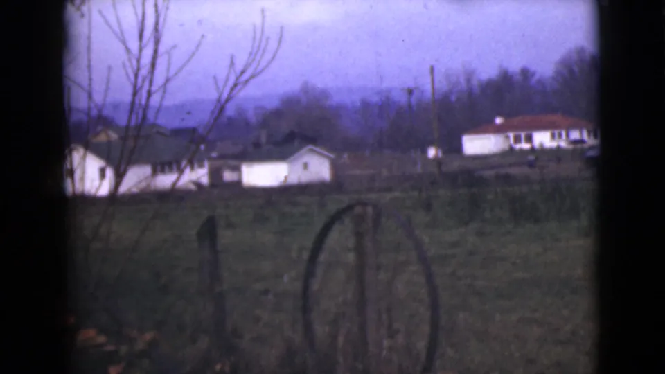 1946 Alaska home-movie view of a rural property, with white outbuildings, a weathered fence, open land, and distant hills.