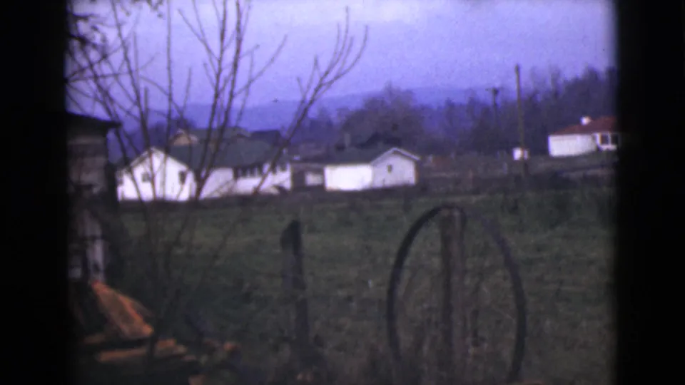 1946 Alaska landscape frame showing a rural yard, white buildings, weathered fence posts, bare tree limbs, and mountains in the distance.