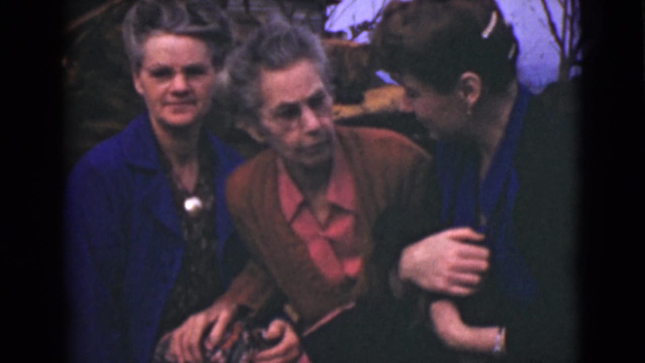Three older women sit close together outdoors, with one leaning toward another.