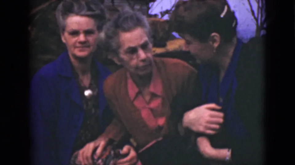 Three older women in 1946 Alaska home-movie footage sit close outdoors, one in a blue coat and another in a brown cardigan with a pink blouse.