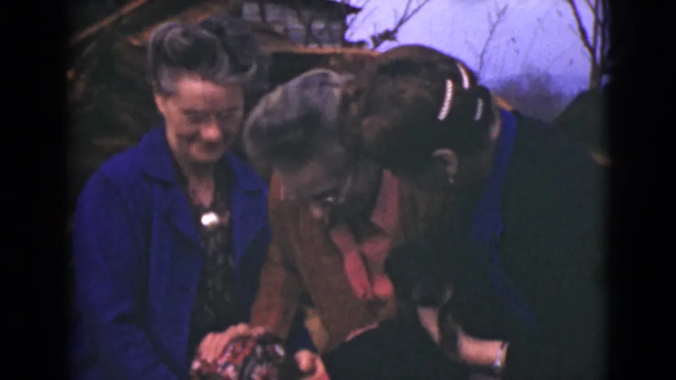 Three older women in 1946 Alaska footage bend forward together, laughing and holding close in a candid outdoor home-movie moment.