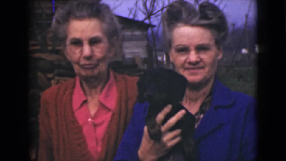 Two older women in 1946 Alaska footage stand outdoors, with the woman in blue holding a small black puppy or animal.
