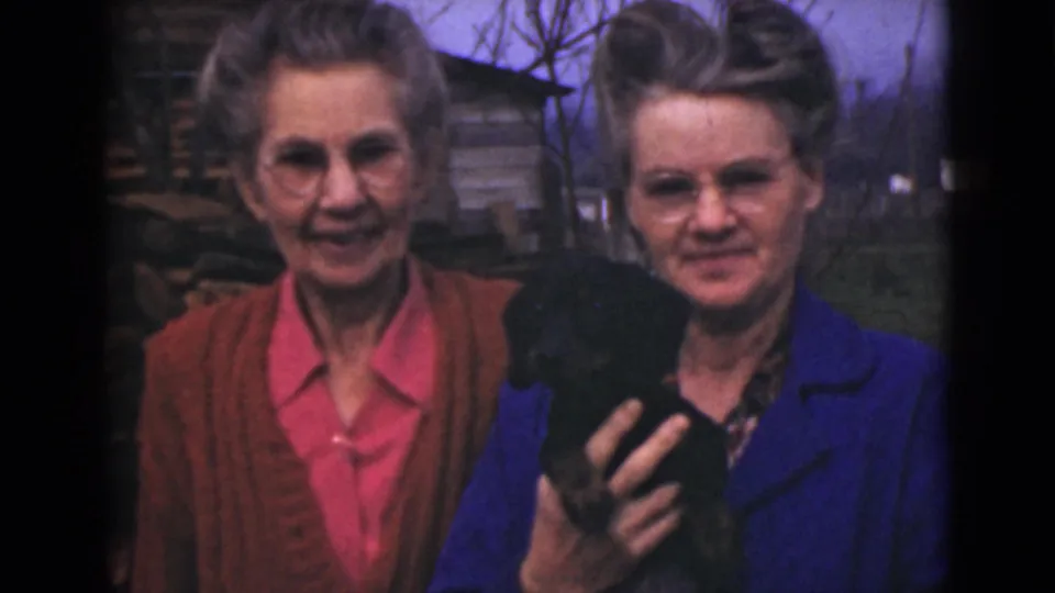 Soft 8mm frame from Alaska in 1946 showing two women outdoors, smiling as one cradles a small black puppy or animal.