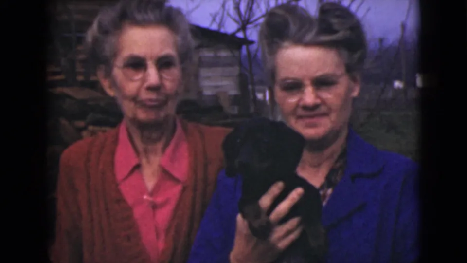 1946 Alaska home-movie still of two older women standing close outdoors as one holds a small black puppy or animal.