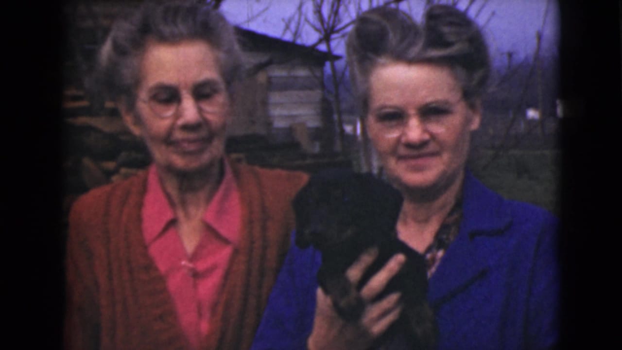 Two women in 1946 Alaska home-movie footage stand outdoors near rough wooden structures, with one holding a small black dog.
