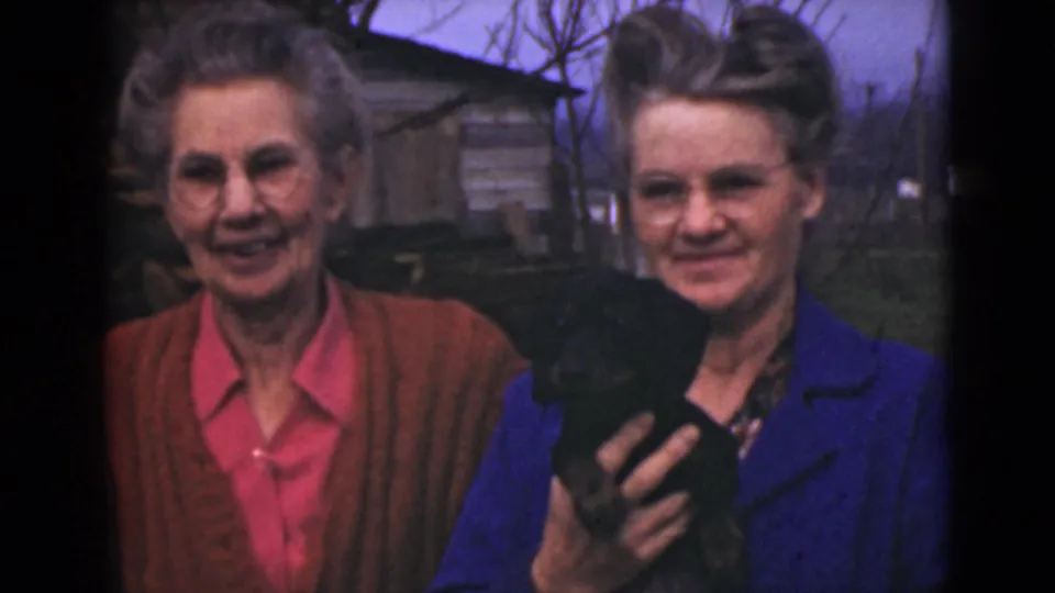 In a 1946 Alaska outdoor home-movie frame, two women look toward the camera while one holds a small black dog close to her chest.