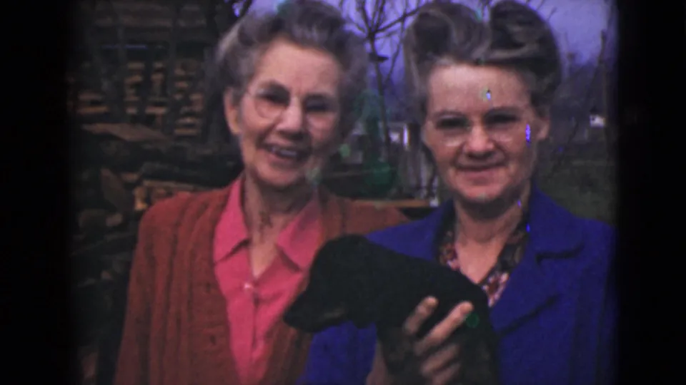 Two women smile in a 1946 Alaska home-movie frame, with a small black dog partly visible in the foreground.