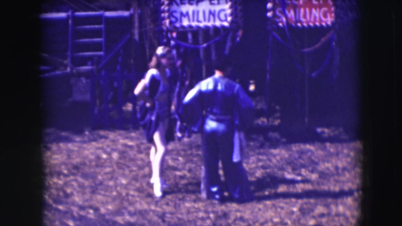 Costumed woman and man in dark uniform stand in a dirt circus area near KEEP EM SMILING signs.