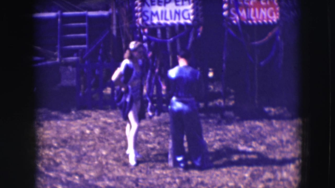 1948 8mm circus-area frame showing a woman in costume and a man facing away under partially readable KEEP EM SMILING signs.