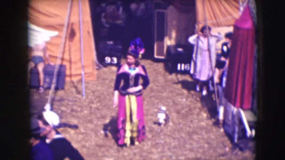 1948 8mm frame showing a costumed woman crossing a Sarasota circus or fairground area with tents and numbered props nearby.
