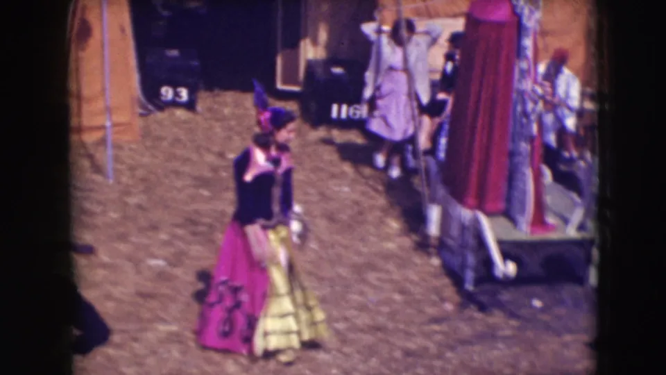 1948 8mm circus-frame detail showing a costumed woman moving past tented structures and workers in the fairground area.