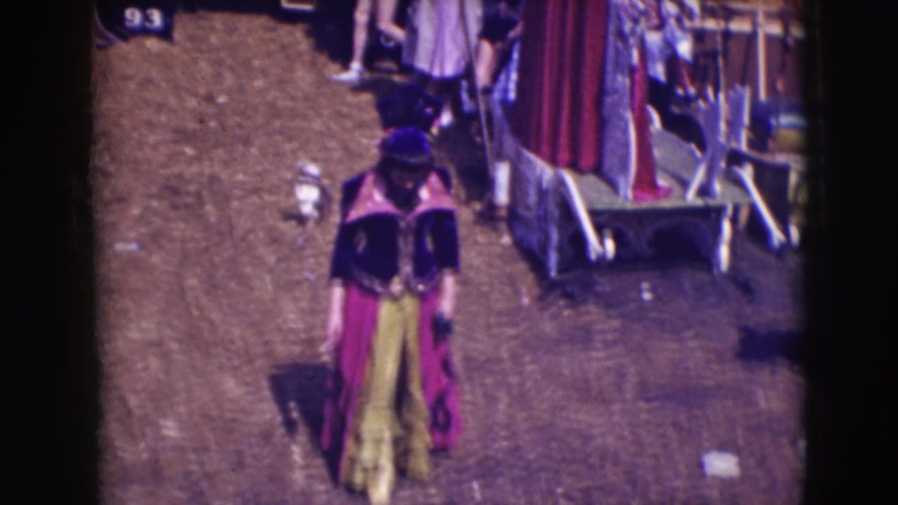 Costumed performer moves through the Ringling Brothers Sarasota backstage area with wagons and people behind them.