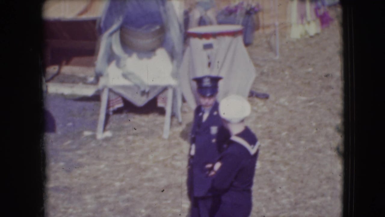 Two uniformed men stand on a dirt circus lot near folding chairs and costumed figures.