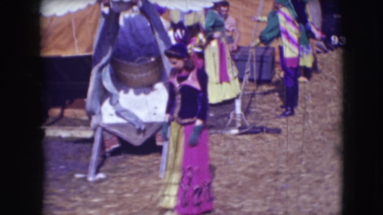Colorfully dressed circus performers stand near tent supports and a large prop or animal covering.