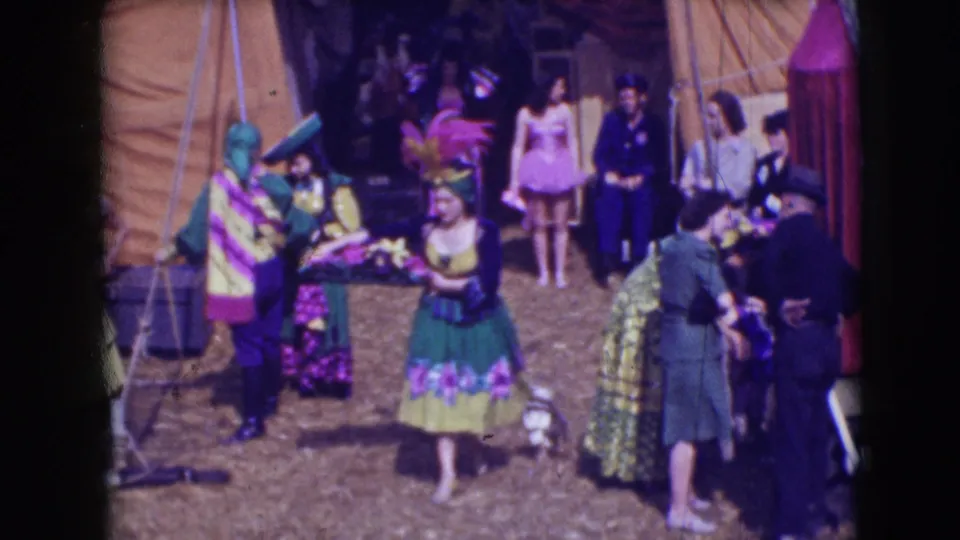 A woman in a green and yellow circus costume carries flowers through the Sarasota tent area while other adults and performers stand nearby.