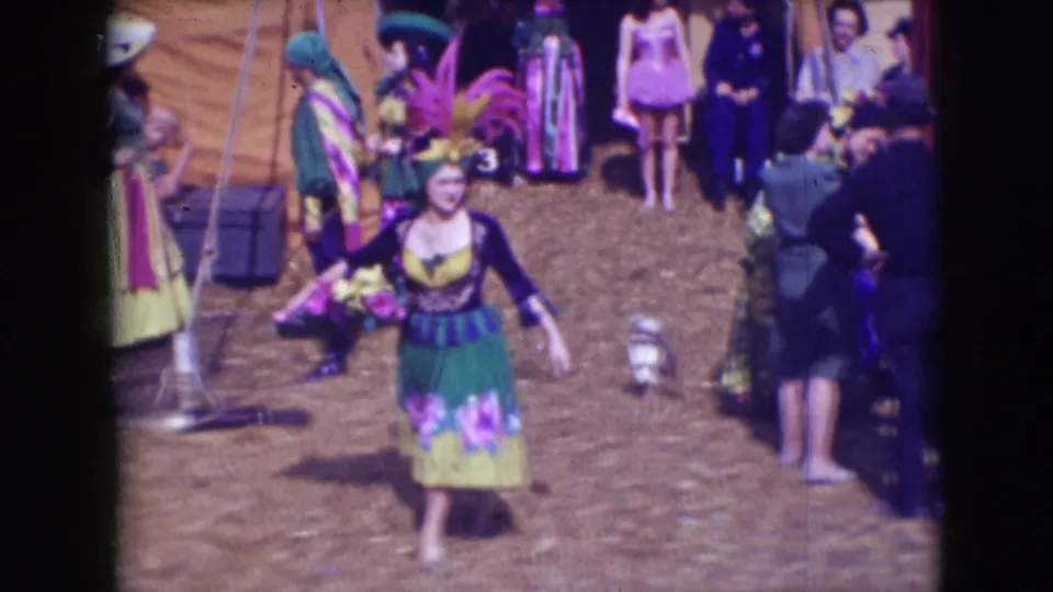 In the 1948 Sarasota circus footage, a performer in a green skirt and flowered costume walks toward camera while pink-clad dancers and staff remain behind.