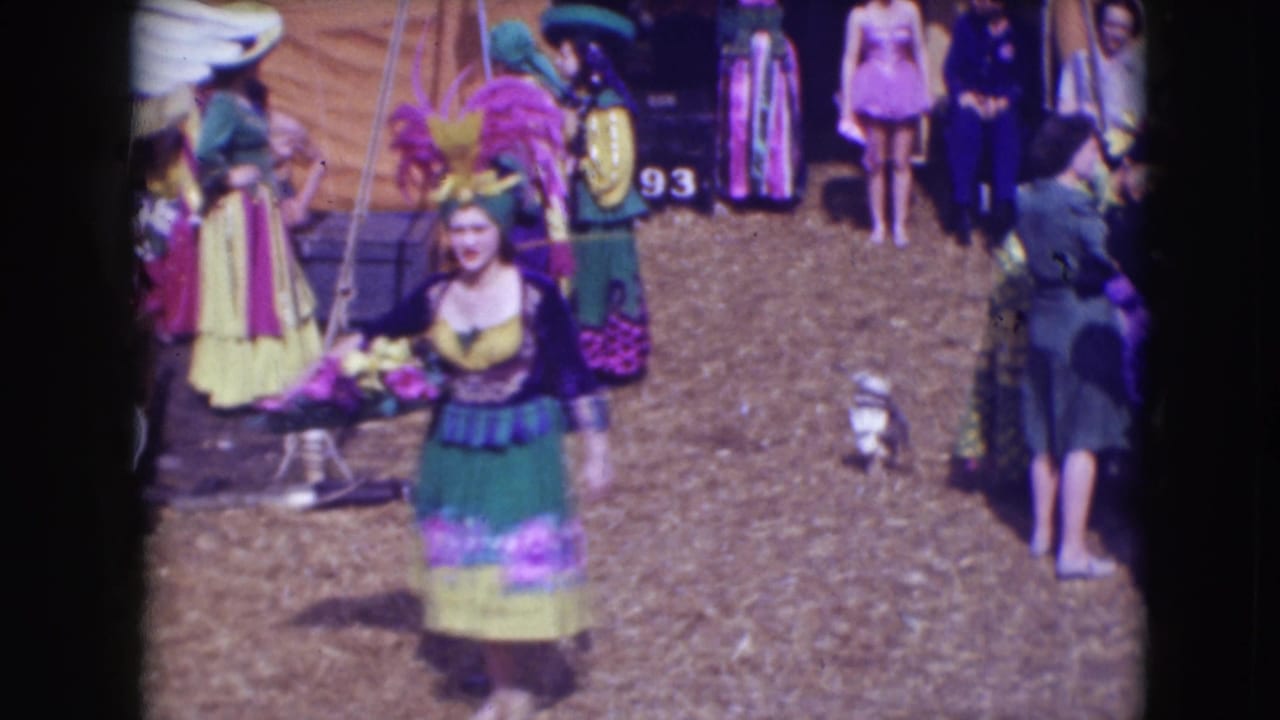 Female circus performer in a green and yellow costume walks across a dirt area near other performers.