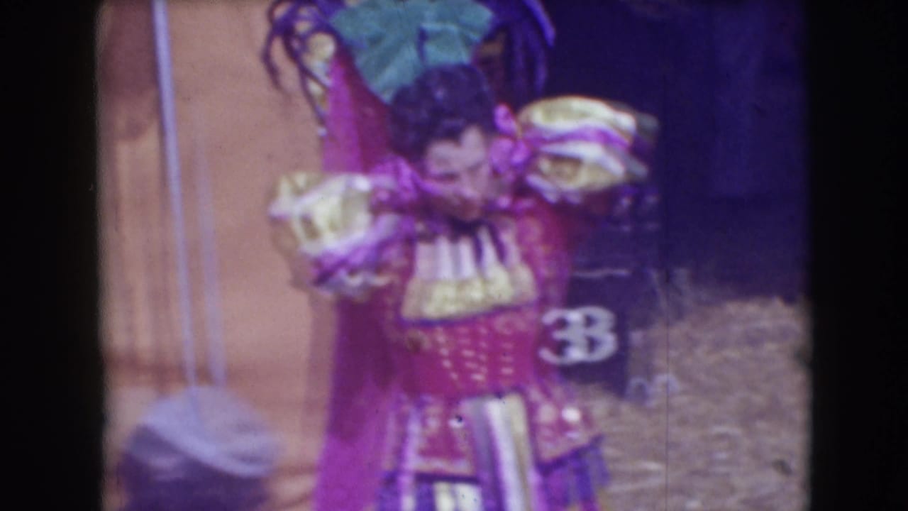 Costumed male performer stands near tent canvas with feathered costume pieces behind him.