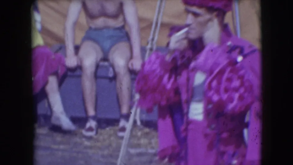 The purple-costumed performer smokes beside seated people and metal supports in a candid 1948 Sarasota circus backstage view.